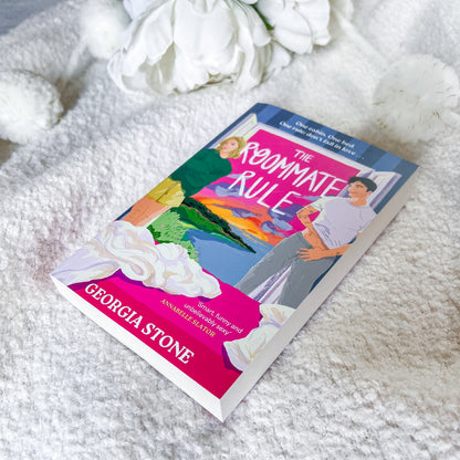 A paperback book titled "The Roommate Rule" by Georgia Stone lies on a soft white textured blanket with white flowers in the background. The cover features a colorful illustration of a couple with bright pink, blue, green, and orange tones.