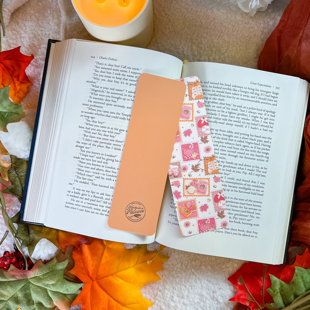 Two decorative book bookmarks displayed on an open book, surrounded by autumn leaves and a candle. One bookmark is solid peach, and the other features a colorful floral and patterned design.