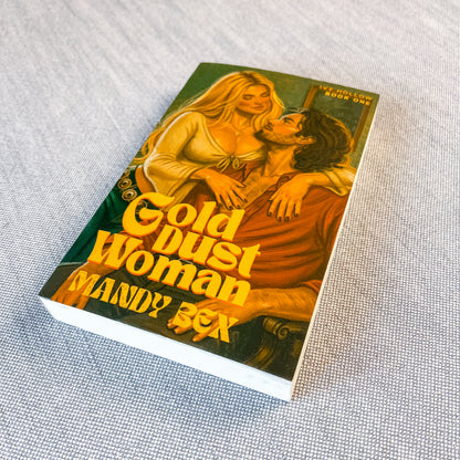 A single paperback book titled "Gold Dust Woman" by Mandy Box, photographed at an angle on a textured light gray fabric surface. The cover features illustrated artwork of a blonde woman in a cream dress embracing a dark-haired man in a rust-colored robe, with large yellow title text across the front.