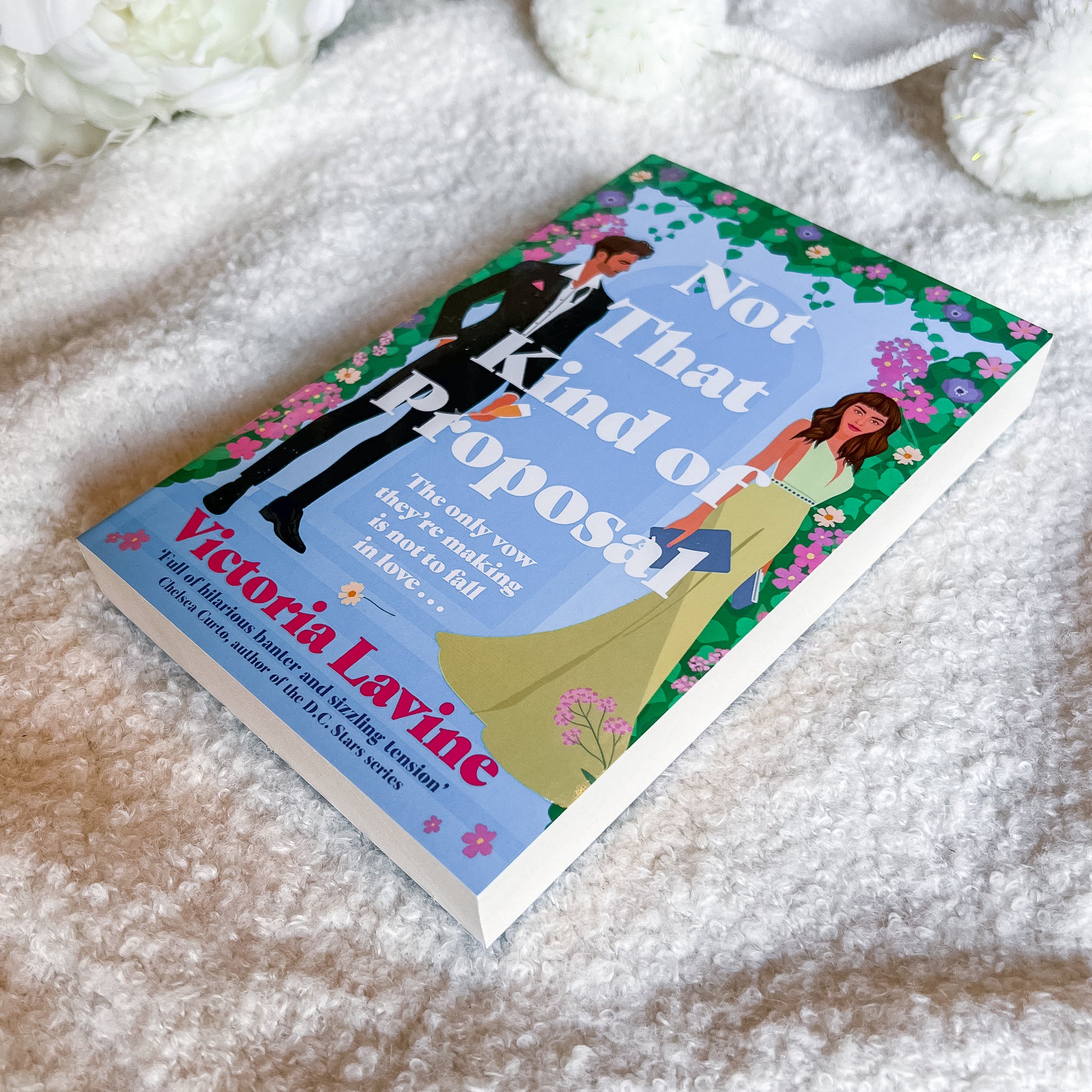 A hardcover book titled "Not That Kind of Proposal" by Victoria Layne, featuring a colorful cover with a man in a black suit and a woman in a yellow-green dress, photographed on a soft white textured blanket with white flowers in the background.