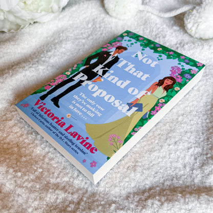 A hardcover book titled "Not That Kind of Proposal" by Victoria Layne, featuring a colorful cover with a man in a black suit and a woman in a yellow-green dress, photographed on a soft white textured blanket with white flowers in the background.