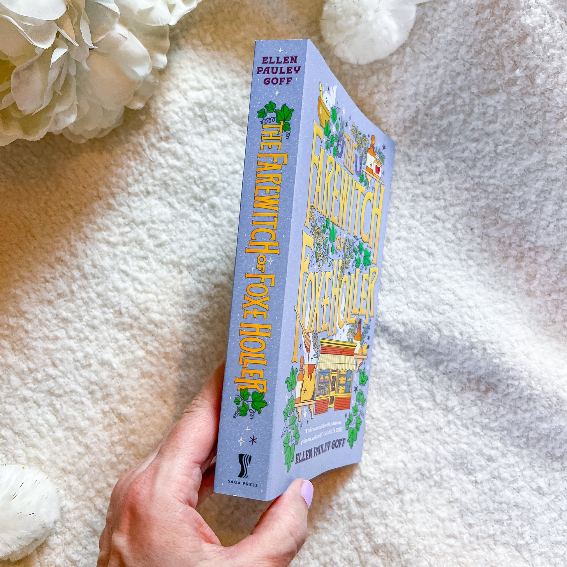 A hand holds a paperback book titled  by Ellen Pavlev Goff, featuring a lavender cover with yellow title text and green clover illustrations, set against a soft white textured blanket with white flowers.