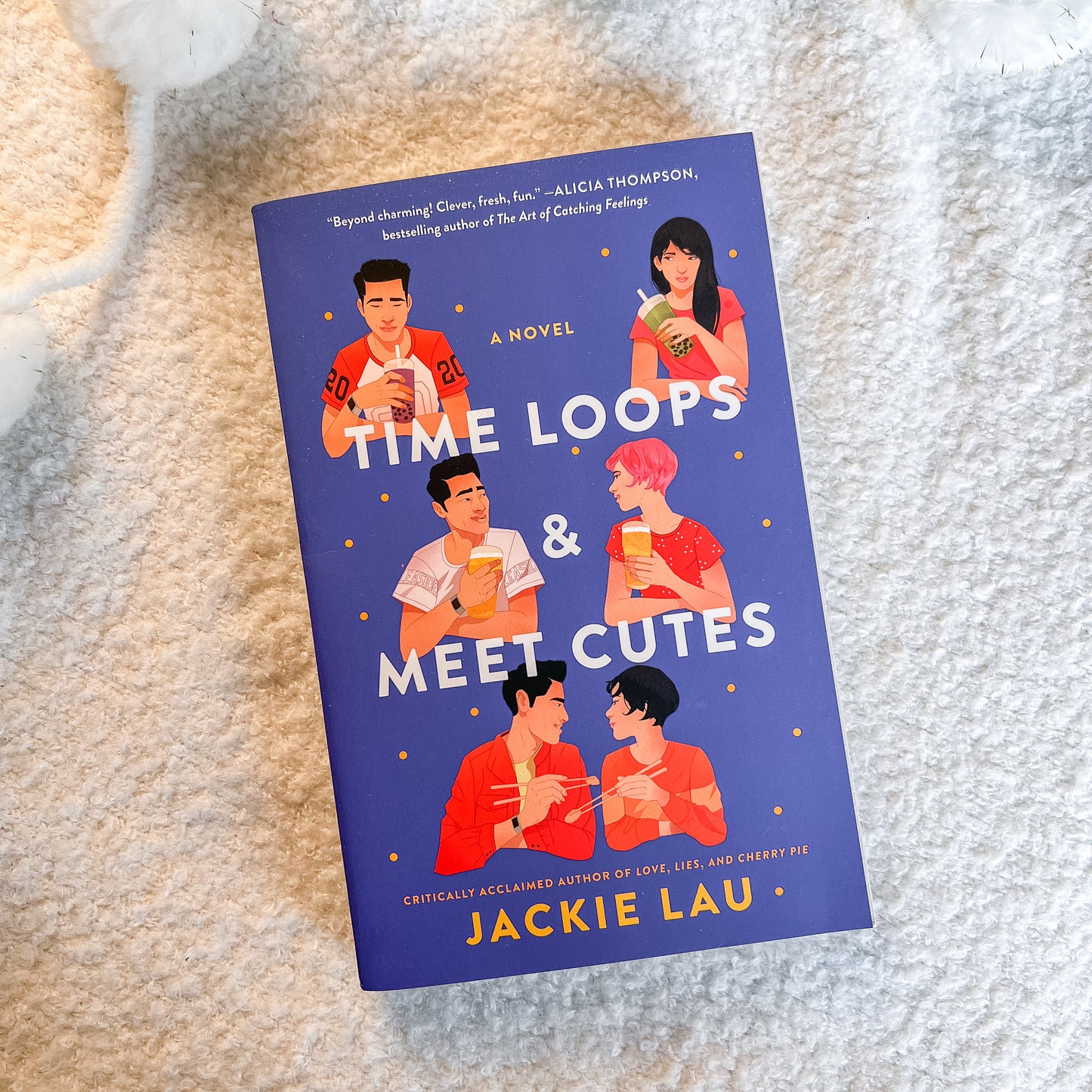 The book cover of the novel 'Time Loops & Meet Cutes' by Jackie Lau, featuring a dark blue background with illustrations of young adults and white title text, photographed on a soft beige textured blanket.