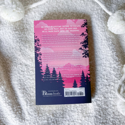 The back cover of a paperback book titled 'He Spent a Lifetime Trying to Escape His Own Past, But to Save Her, He'll Wade Back Into the Shadows' by Bloom Books, placed on a textured white blanket.