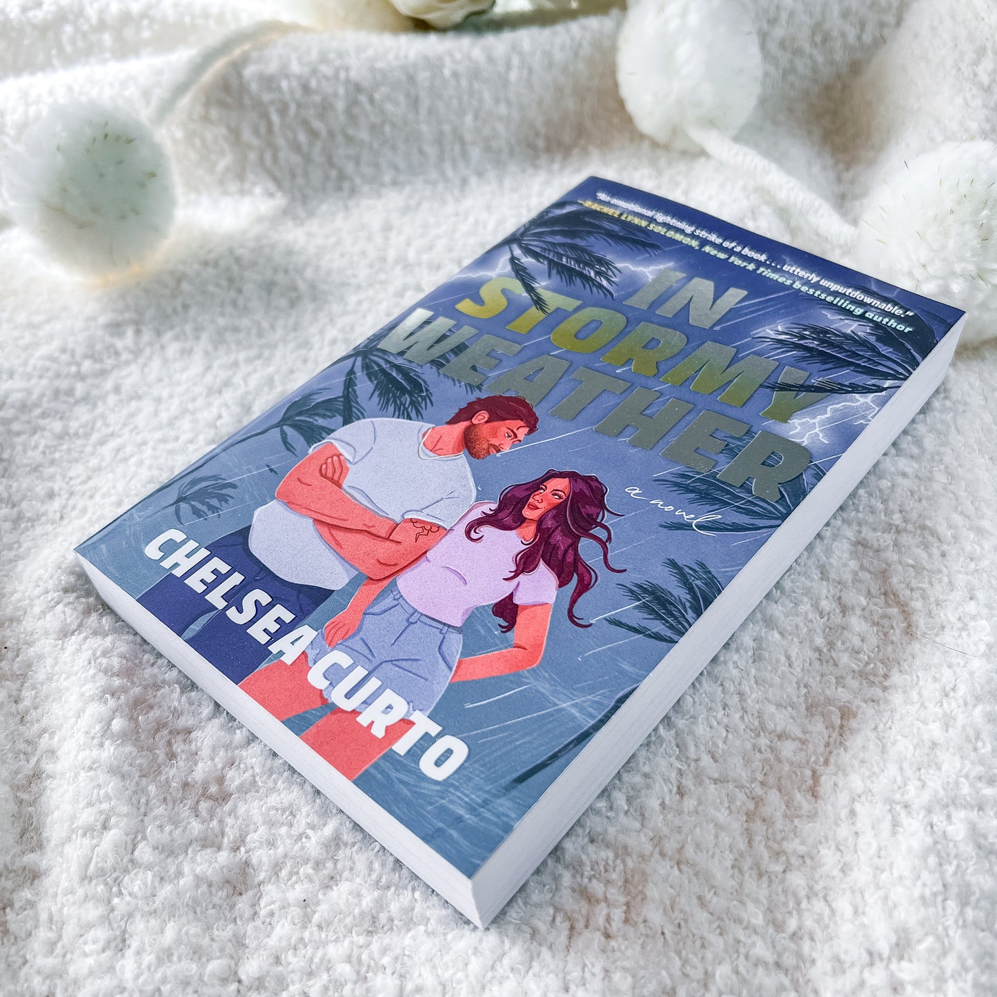 A paperback copy of the book 'In Stormy Weather' by Chelsea Curto, featuring a blue-toned tropical illustration of a couple, displayed on a soft white textured blanket.