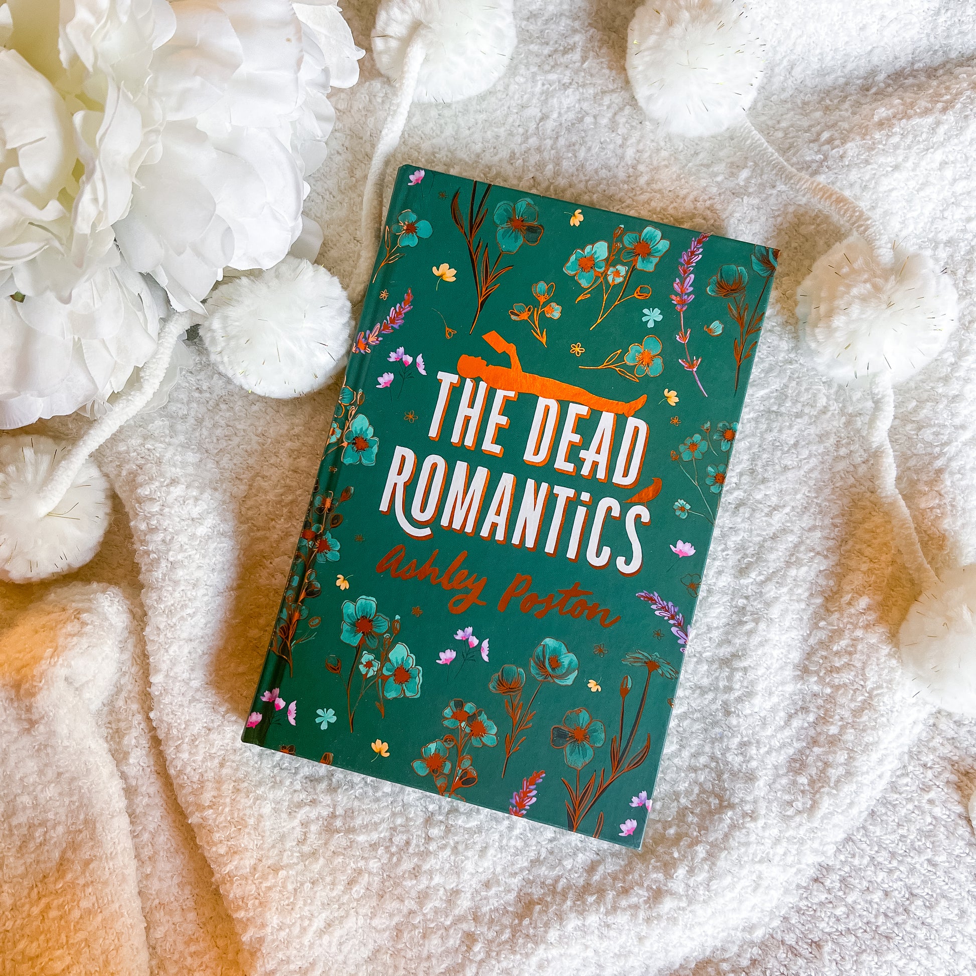Book titled 'The Dead Romantics' by Ashley Poston on a soft fabric background with flowers.