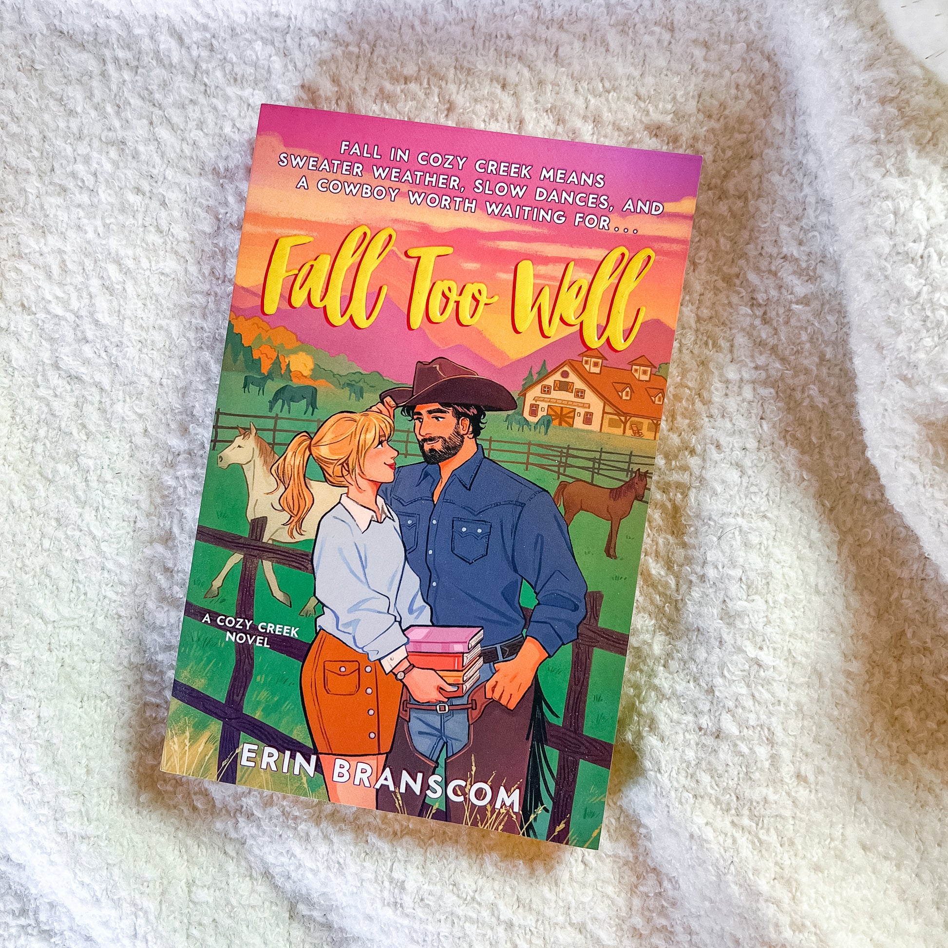 A book titled "Fall Too Well" by Erin Branscom, featuring a colorful illustrated cover with a cowboy and cowgirl in a farm setting, placed on a white textured blanket.