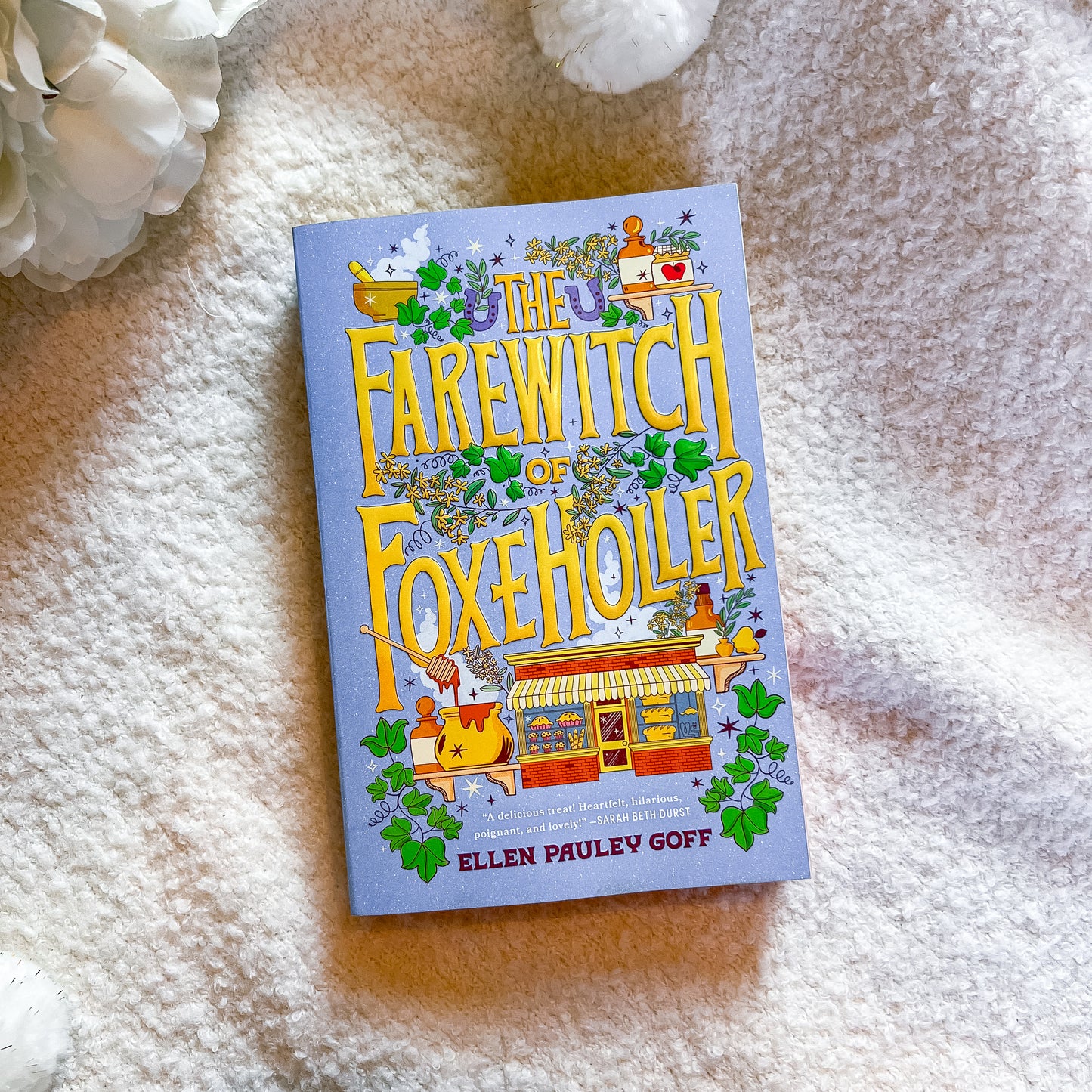 The book 'The Farewitch of Foxe Holler' by Ellen Pauley Goff, featuring a light blue cover with whimsical illustrations and large yellow title text, photographed on a soft cream-colored textured blanket with white decorative flowers in the corners.