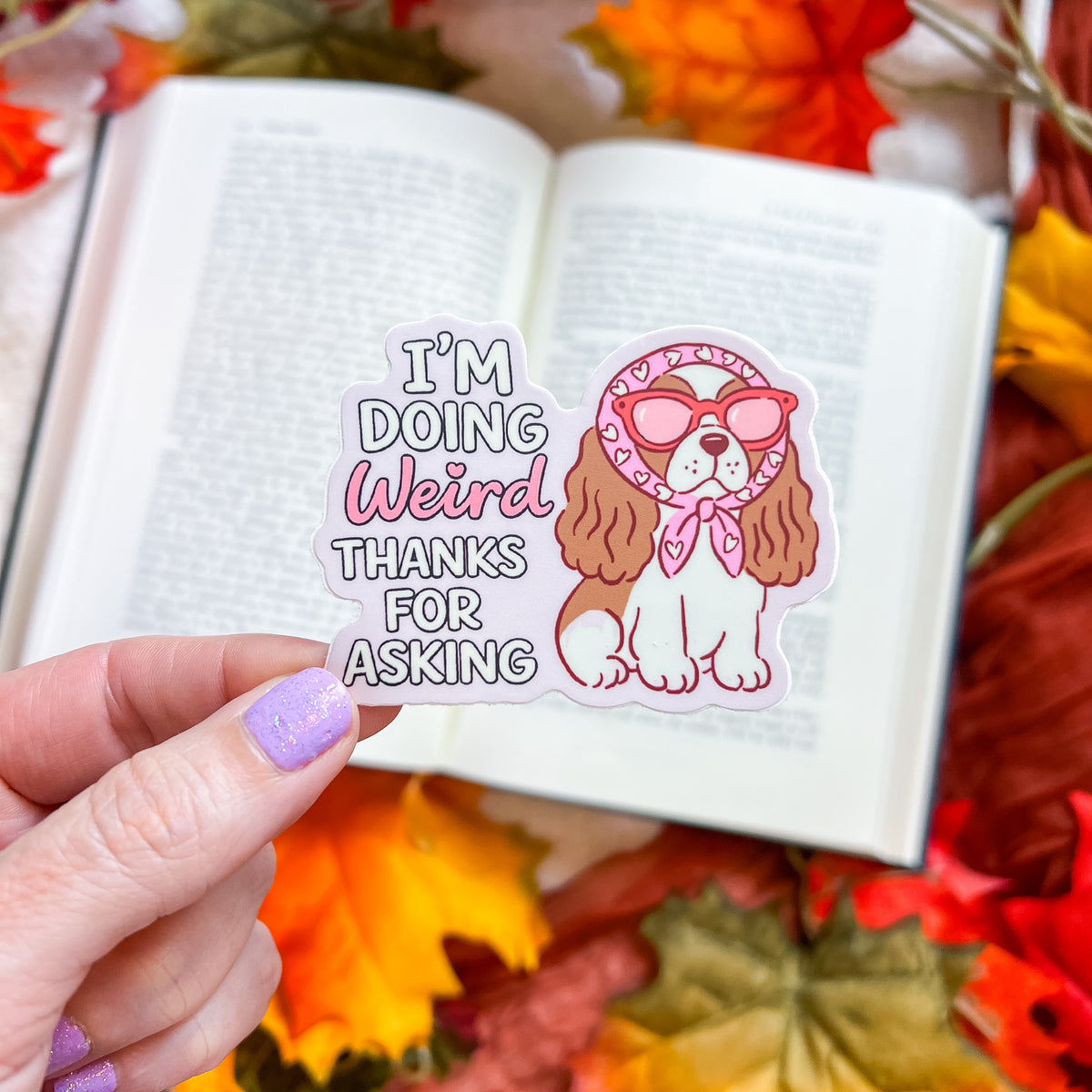 A hand with lavender glitter nail polish holds a die-cut sticker in front of an open book surrounded by autumn leaves. The sticker features a cartoon Cavalier King Charles Spaniel dog wearing pink sunglasses and a pink bow tie, next to the text 'I'M DOING Weird THANKS FOR ASKING'.