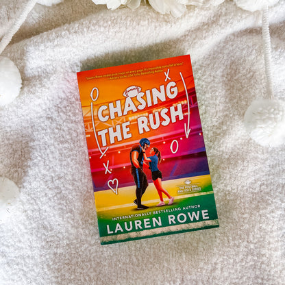 The book 'Chasing the Rush' by Lauren Rowe is displayed flat on a soft, textured white blanket with decorative white flowers in the corners. The book cover features a vibrant, colorful gradient background transitioning from orange and yellow to purple and green, with a graphic illustration of a football player and a woman running together.