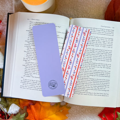 A lavender purple bookmark with a circular logo, placed on an open book next to a decorative bookmark featuring an orange and purple pattern.