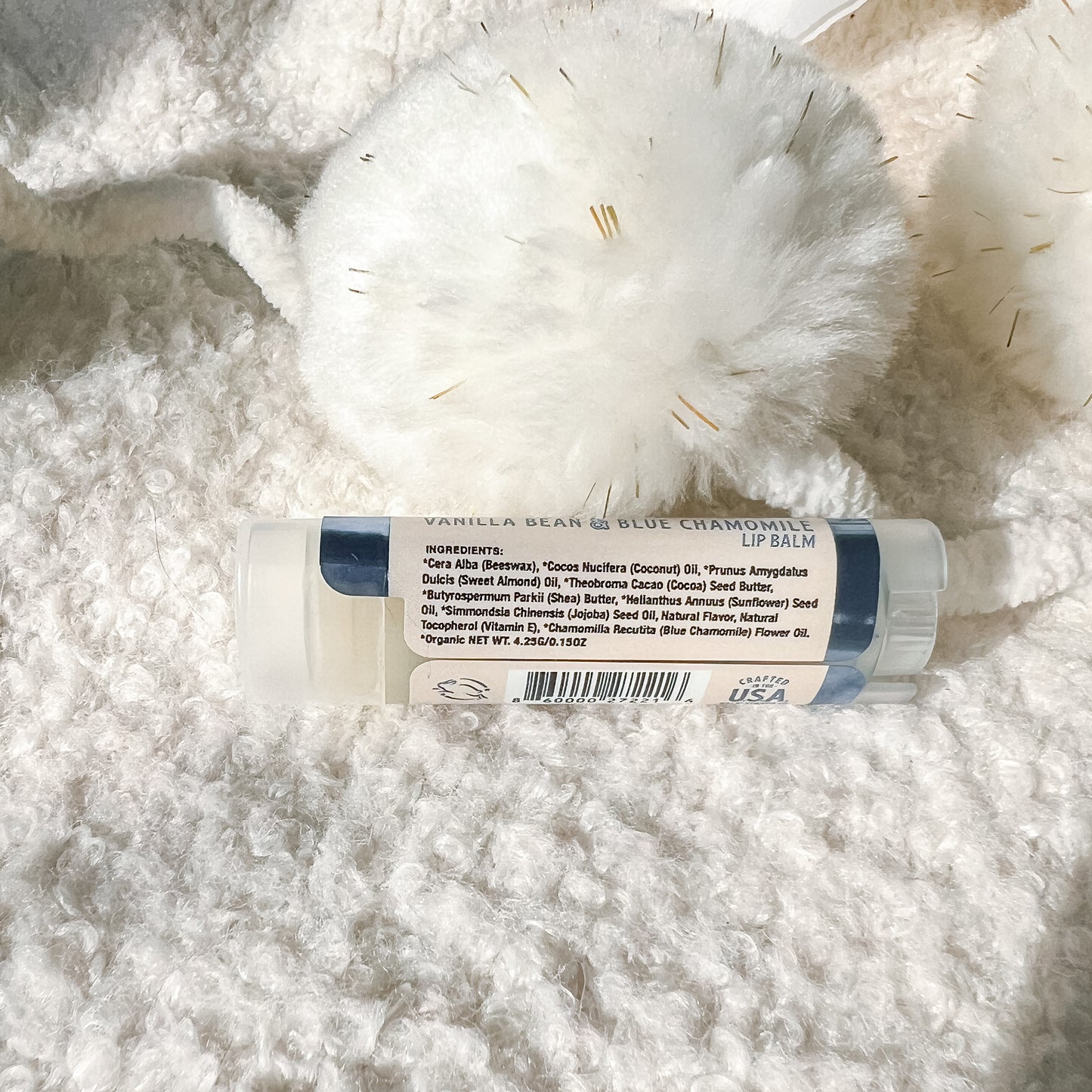 A lip balm tube labeled 'Vanilla Bean & Blue Chamomile Lip Balm' resting on a white textured faux fur surface next to a white pom-pom.
