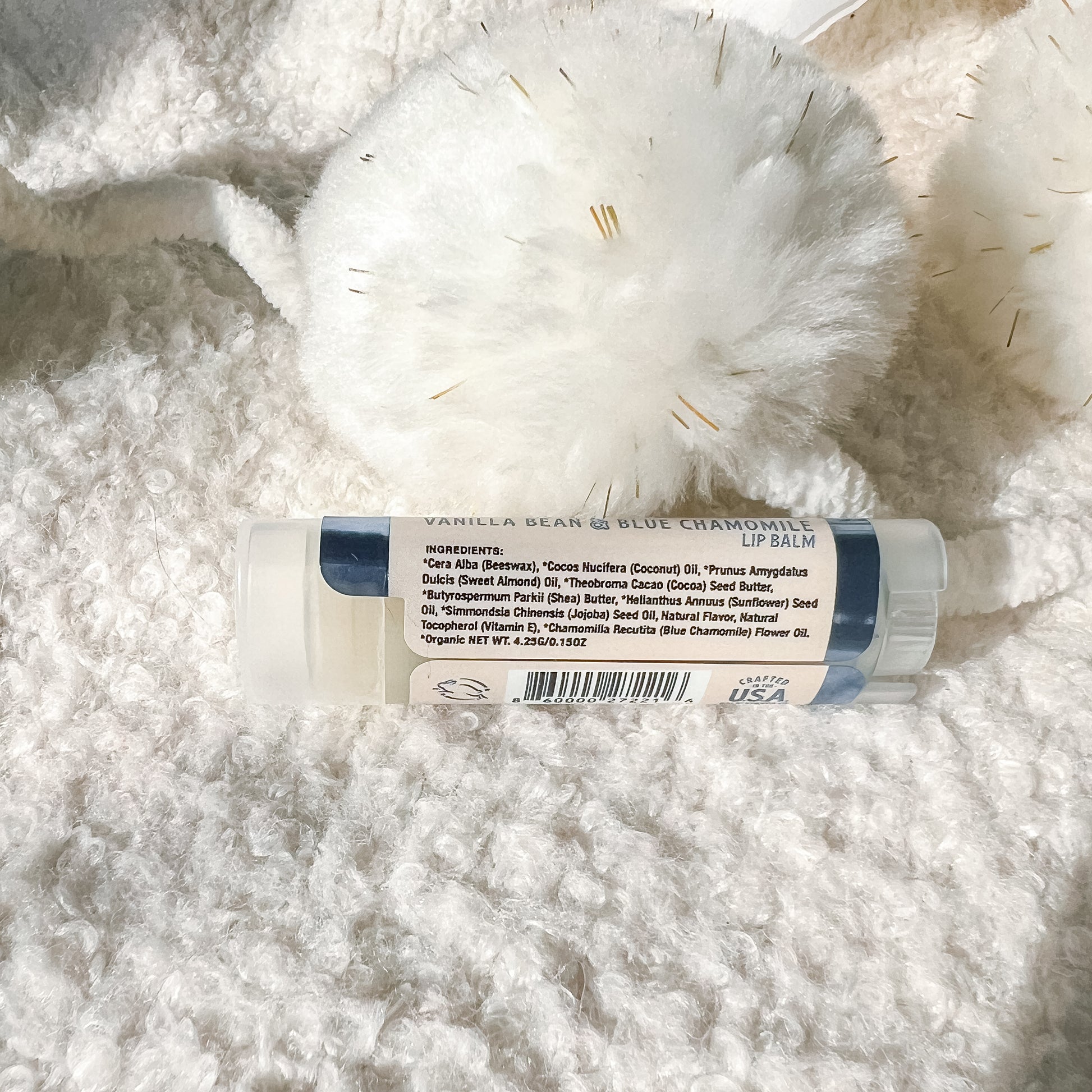 A lip balm tube labeled 'Vanilla Bean & Blue Chamomile Lip Balm' resting on a white textured faux fur surface next to a white pom-pom.