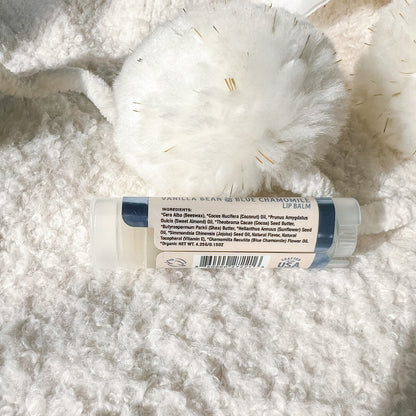 A lip balm tube labeled 'Vanilla Bean & Blue Chamomile Lip Balm' resting on a white textured faux fur surface next to a white pom-pom.