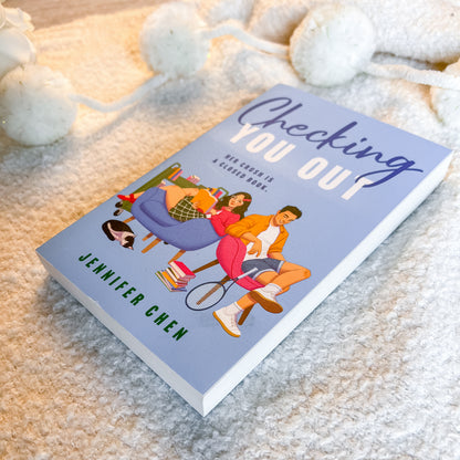 A paperback book titled 'Checking You Out' by Jennifer Chen lies on a soft beige textured blanket with white pom-pom garlands in the background. The cover is light blue with a colorful illustration of a couple sitting on a couch and a tennis racket.