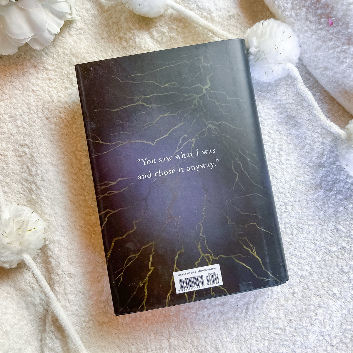 A hardcover book with a dark, textured cover featuring gold vein-like patterns and a quote printed in white, styled on a soft cream-colored blanket with white decorative pom-pom flowers.