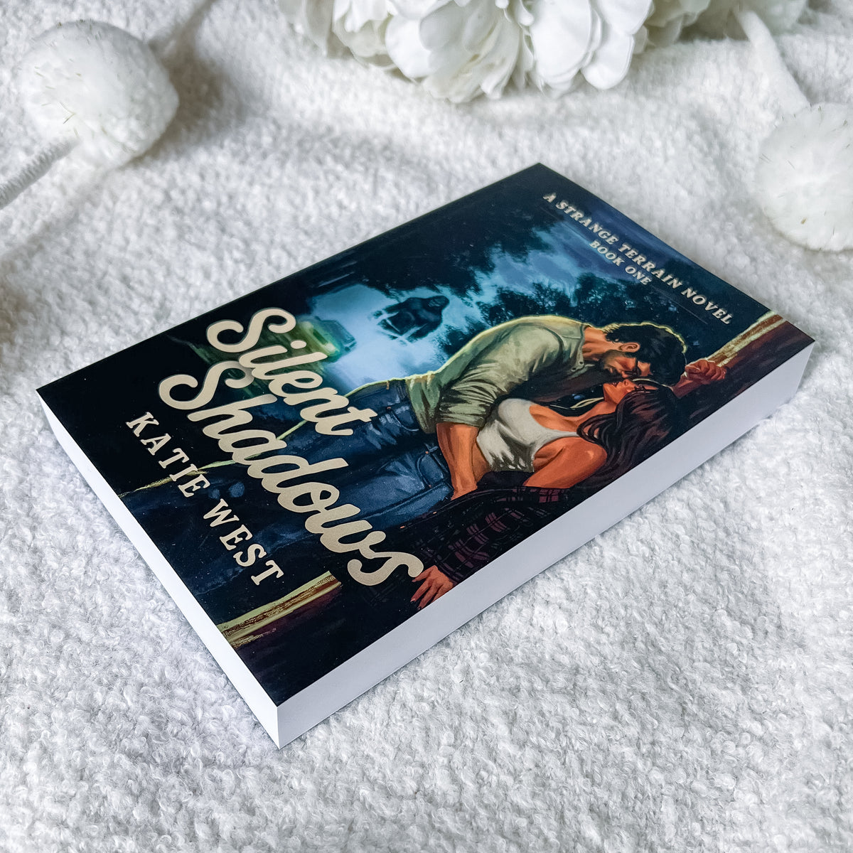 A paperback copy of the novel 'Silent Shadows' by Kate West is displayed on a soft white textured blanket with decorative white flowers in the background. The book cover features a dark, romantic illustration of a couple embracing in a moonlit scene.