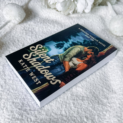 A paperback copy of the novel 'Silent Shadows' by Kate West is displayed on a soft white textured blanket with decorative white flowers in the background. The book cover features a dark, romantic illustration of a couple embracing in a moonlit scene.