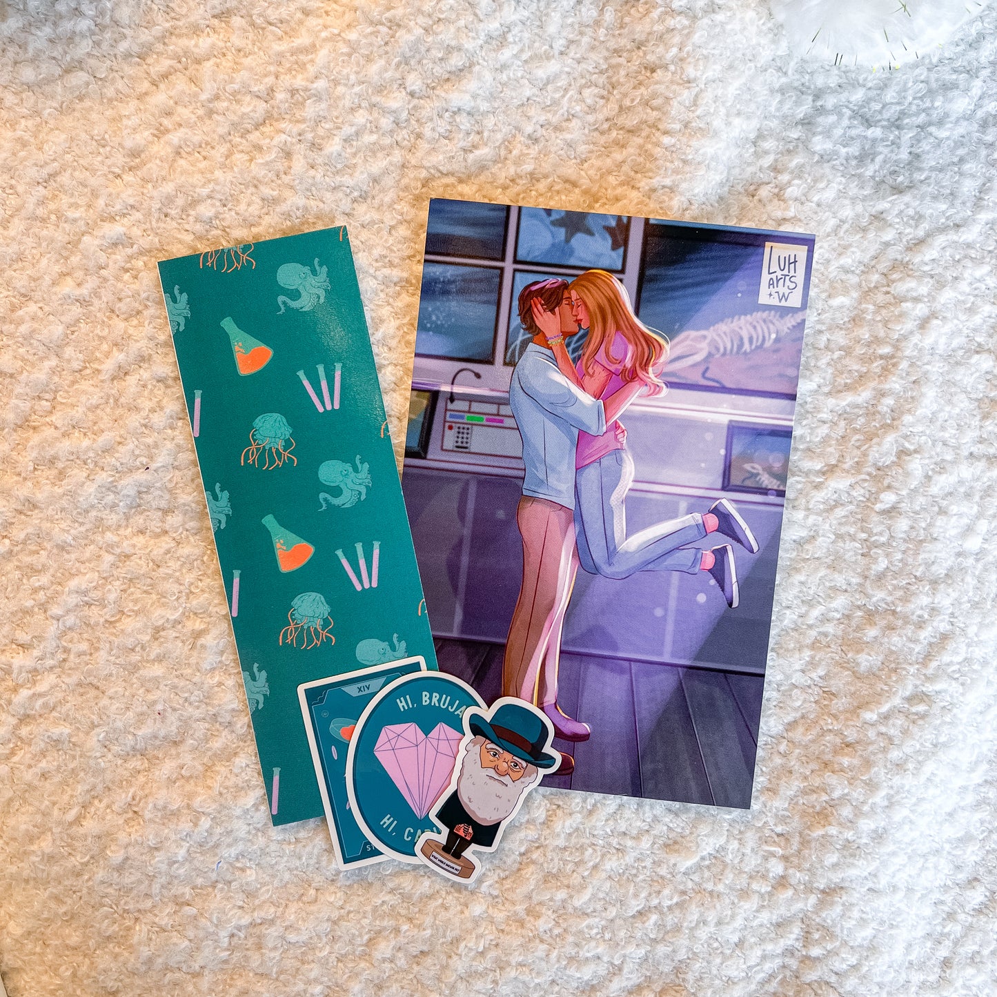 A flat-lay of three collectible items on a textured cream-colored faux fur surface: a teal patterned bookmark, a printed art card featuring an anime-style couple, and two die-cut stickers with cartoon characters and text.