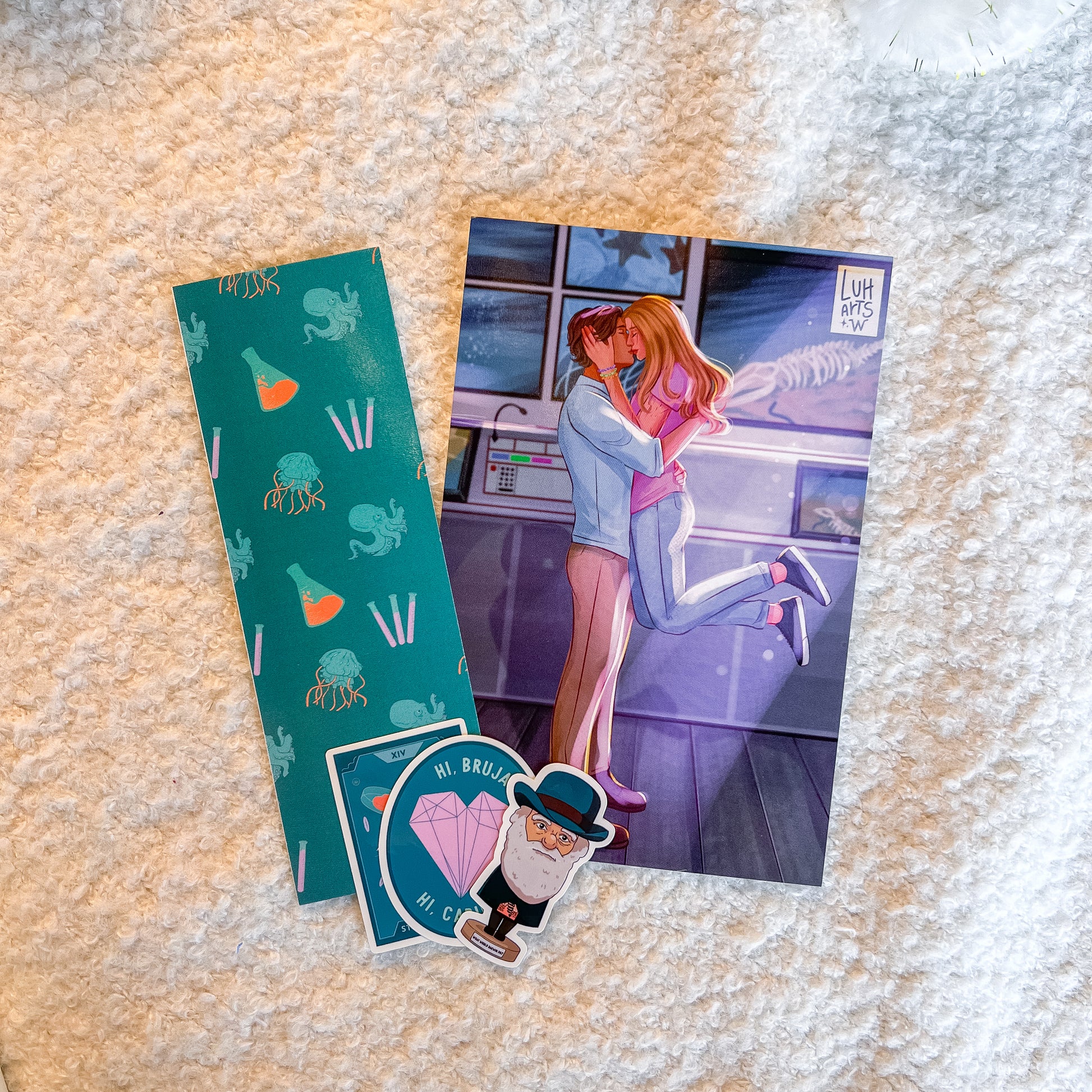A flat-lay of three collectible items on a textured cream-colored faux fur surface: a teal patterned bookmark, a printed art card featuring an anime-style couple, and two die-cut stickers with cartoon characters and text.