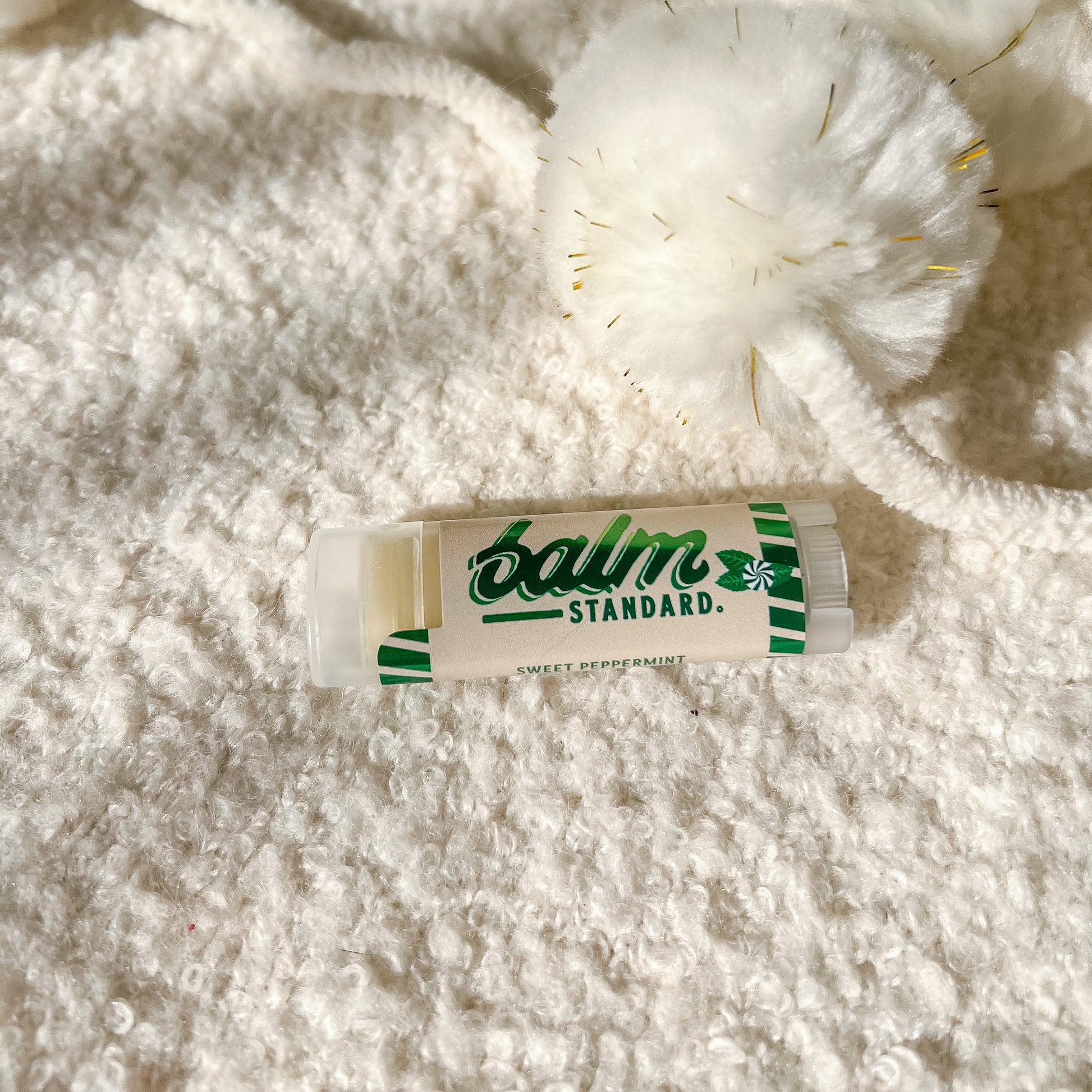 A tube of Balm Standard Sweet Peppermint lip balm lies on a white textured blanket next to a white pom-pom with gold thread details.