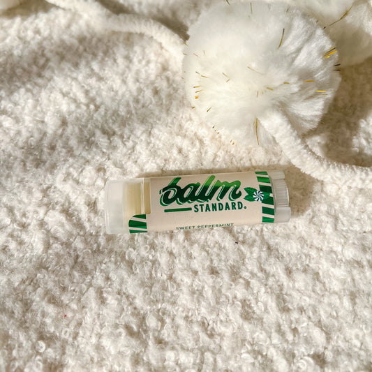 A tube of Balm Standard Sweet Peppermint lip balm lies on a white textured blanket next to a white pom-pom with gold thread details.