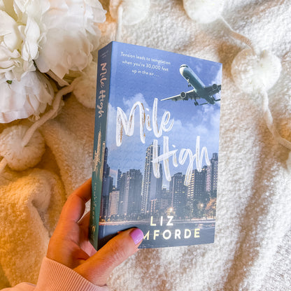 Book titled 'Mile High' by Liz Amforde held against a soft, textured background with flowers.