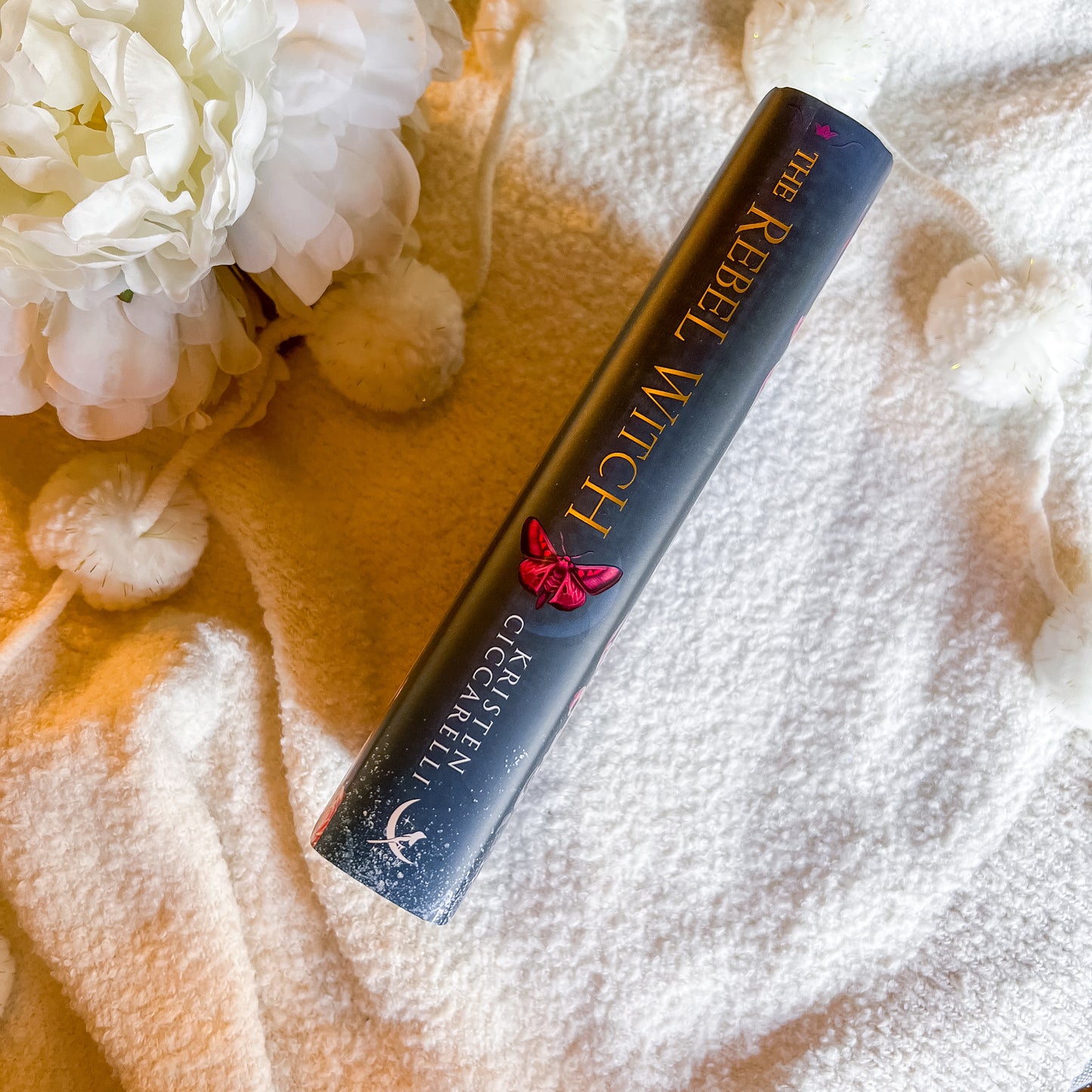 Spine of "The Rebel Witch" book on a textured surface with flowers in the background