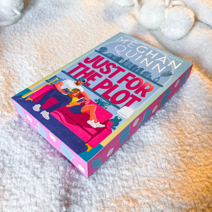 A hardcover book titled 'Just For The Plot' by Meaghan Quinn lies on a soft white textured blanket. The cover features a colorful illustration of a couple lounging on a pink sofa, with the author's name and title clearly visible.