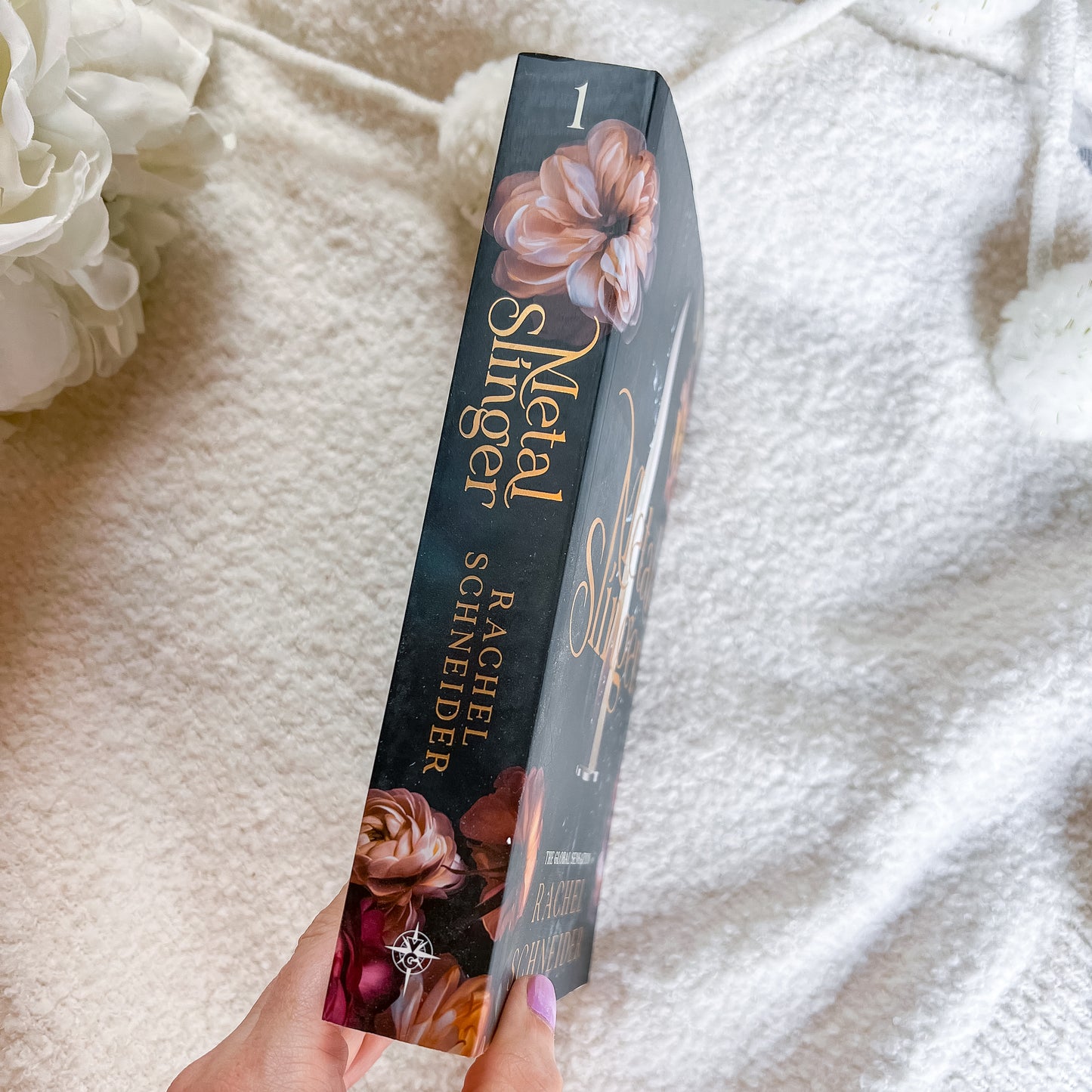 A hand holds a slim book titled 'Metal Slugger' by Rachel Schneider, standing upright against a soft white textured blanket background with white flowers in the corner.