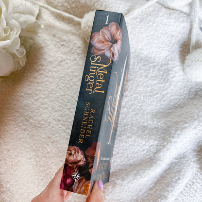 A hand holds a slim book titled 'Metal Slugger' by Rachel Schneider, standing upright against a soft white textured blanket background with white flowers in the corner.