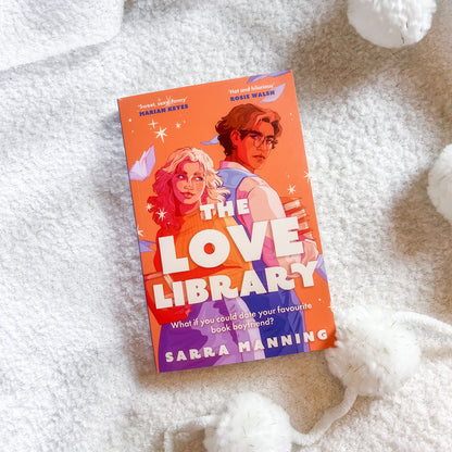 The book cover of 'The Love Library' by Sarra Manning is displayed on a soft, white textured blanket with white pom-pom yarn balls. The cover features a bright orange background with a colorful illustration of a couple and bold white title text.
