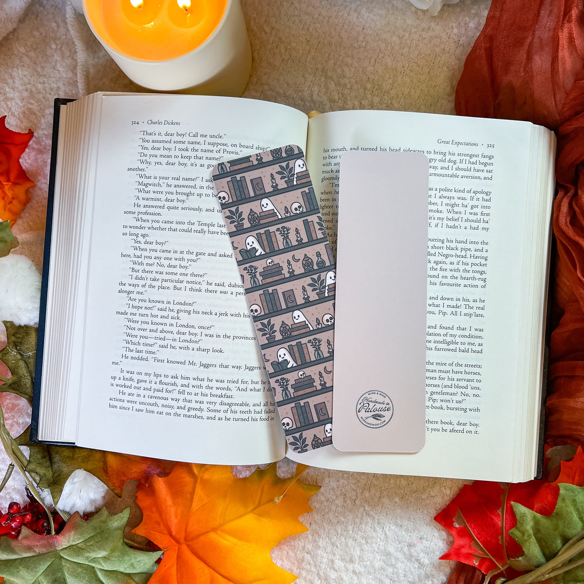 A decorative paper bookmark. The scene is styled with autumn leaves, a candle, and a cozy blanket background.
