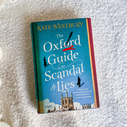 The Oxford Guide to Scandal & Lies novel by Kate Westbury, featuring a teal cover with white title text and a red spine, photographed on a soft white textured blanket.