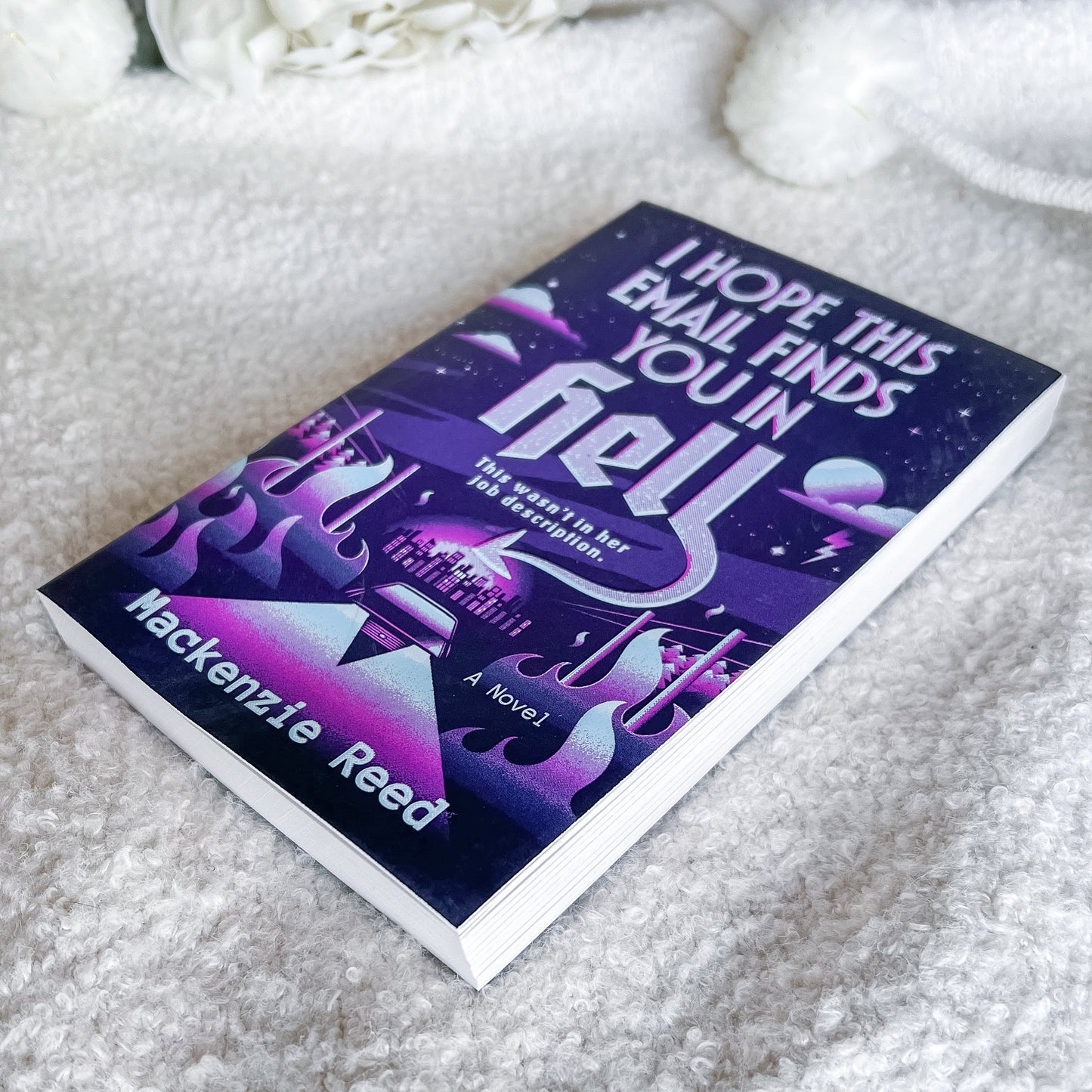 A paperback novel titled "I Hope This Email Finds You in Hell" by Hackenzie Reed, featuring a purple and black cosmic cover with stylized flames, placed on a soft white textured blanket.