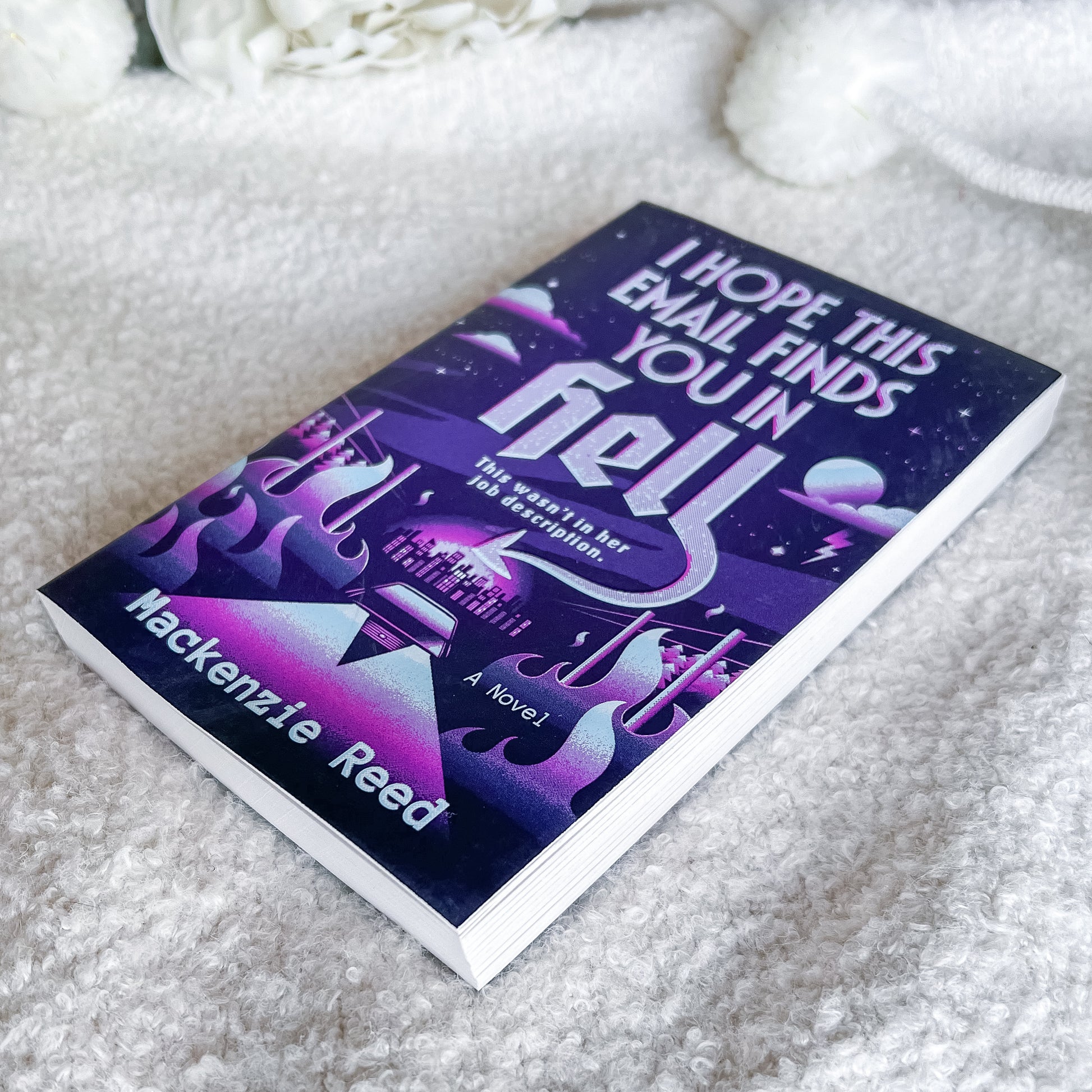 A paperback novel titled "I Hope This Email Finds You in Hell" by Hackenzie Reed, featuring a purple and black cosmic cover with stylized flames, placed on a soft white textured blanket.