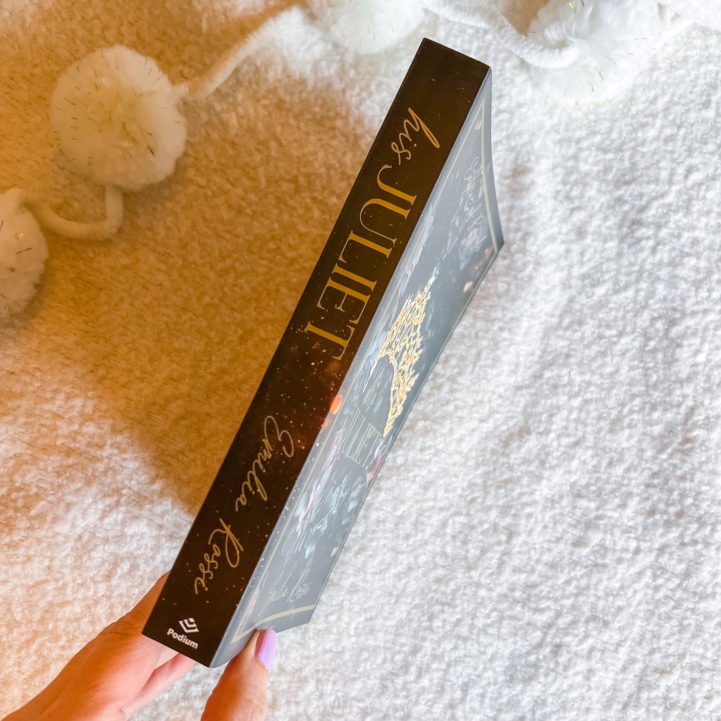 A hand holds a slim book, showing its dark spine with gold lettering, against a soft white textured blanket background with decorative white pom-poms.