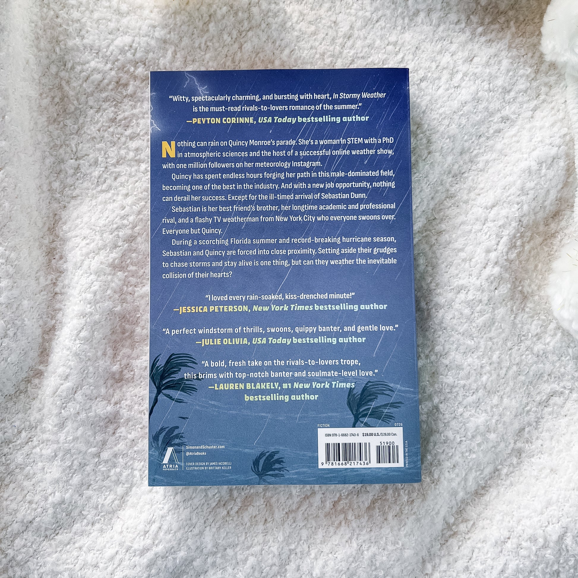 The back cover of a book lying on a white textured blanket. The cover is dark blue with white text, featuring book reviews, a barcode, and tropical palm leaf illustrations at the bottom.