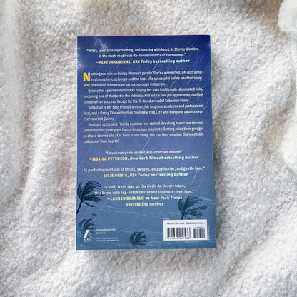 The back cover of a book lying on a white textured blanket. The cover is dark blue with white text, featuring book reviews, a barcode, and tropical palm leaf illustrations at the bottom.
