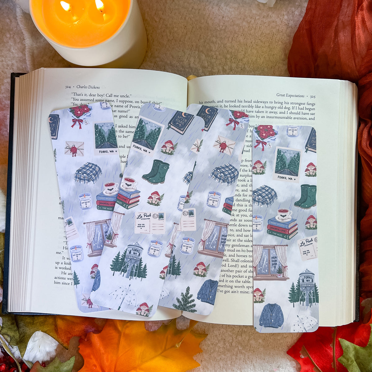 Three rectangular paper bookmarks or tags with a cozy winter cabin illustration print are arranged on an open book, surrounded by autumn leaves, a lit candle, and a warm blanket.