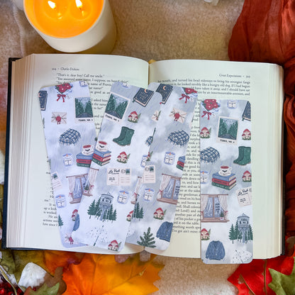Three rectangular paper bookmarks or tags with a cozy winter cabin illustration print are arranged on an open book, surrounded by autumn leaves, a lit candle, and a warm blanket.