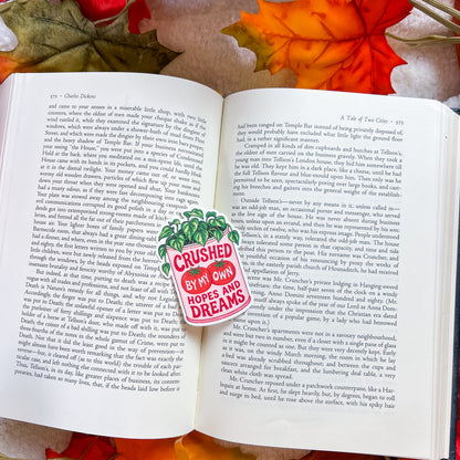 An open book spread photographed from above on a brown textured surface. The left page has black text and a red-and-white sticker reading 'CRUSHED BY MY OWN DREAMS AND DREAMS.