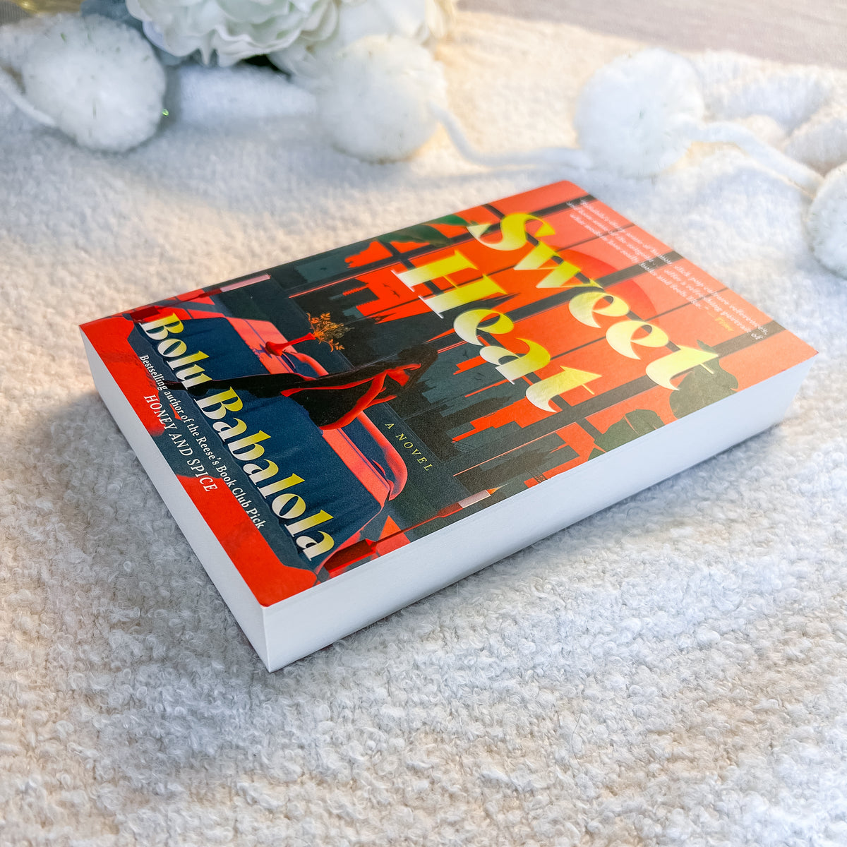 The book 'Sweet Heat' by Boubir Bhalola lies open on a soft, textured cream-colored blanket, with decorative white flowers in the background.