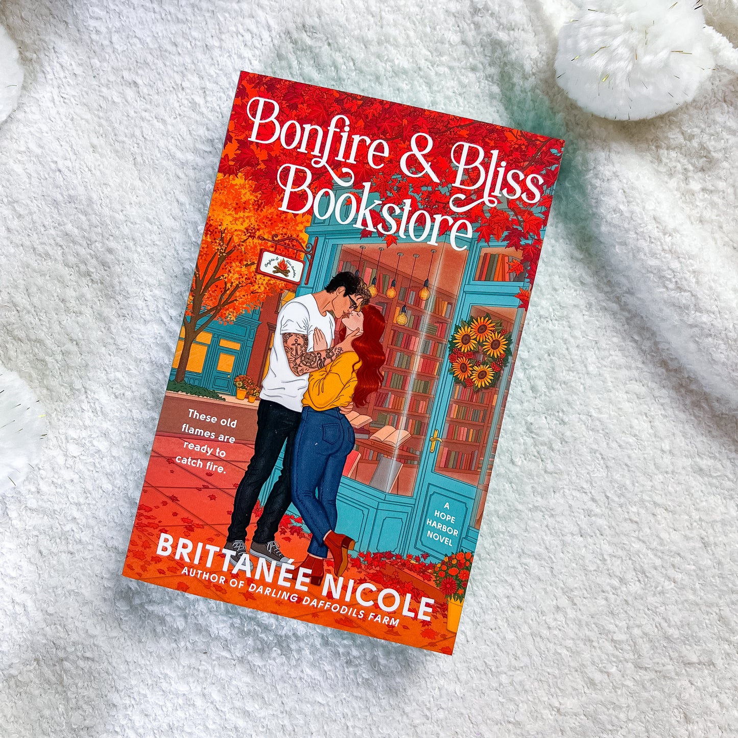 A book titled "Bonfire & Bliss Bookstore" by Brittanée Nicole is photographed flat on a soft white textured blanket. The cover art shows a couple embracing outside a colorful bookstore with autumn leaves and warm orange-red tones.