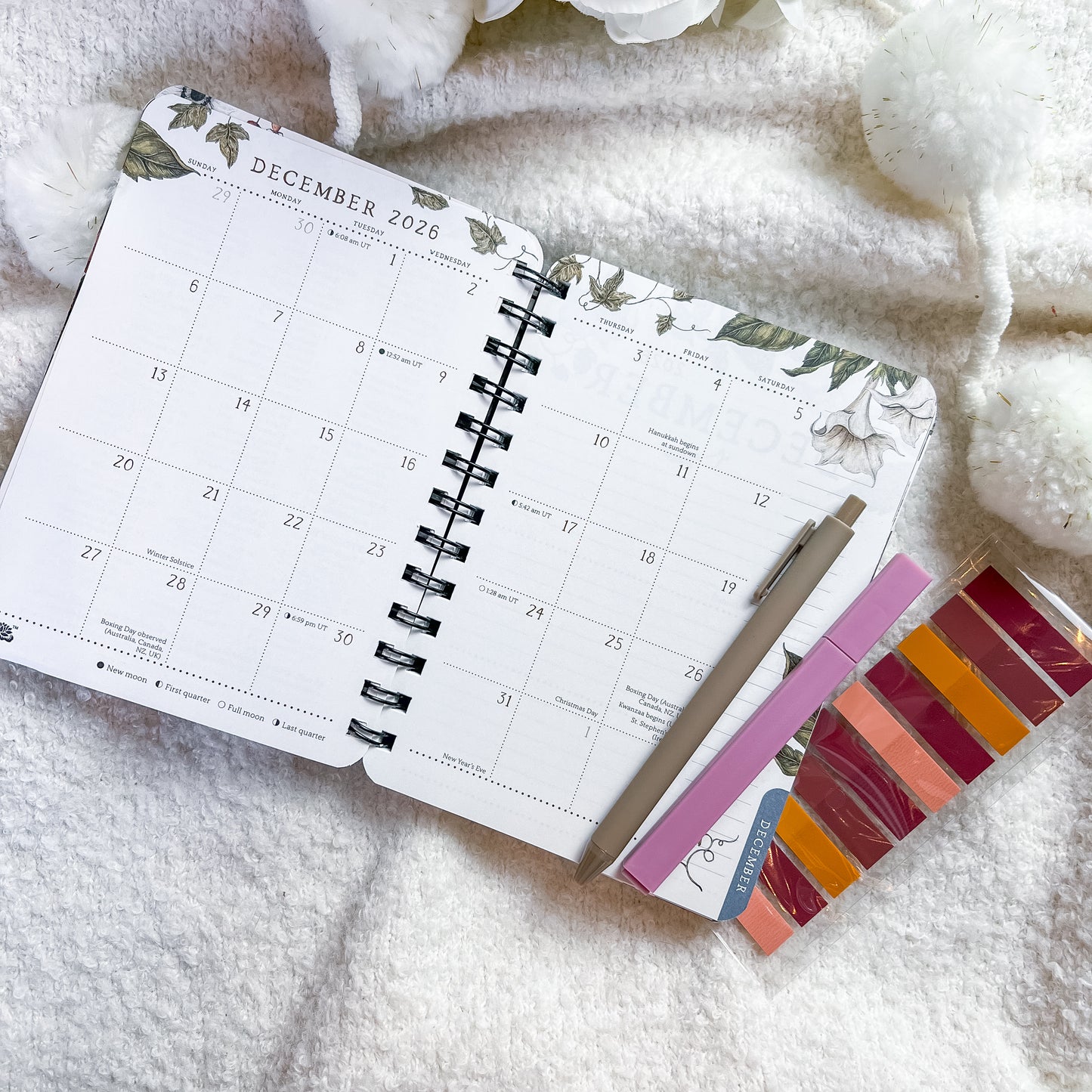 An open spiral-bound planner showing December 2026, styled on a soft white textured blanket with decorative white flowers and a pen. A small card with colorful swatches is tucked under the planner.