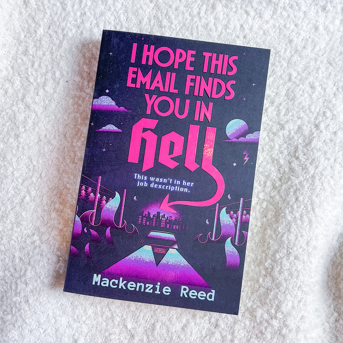 A book titled 'I Hope This Email Finds You In Hell' by Mackenzie Reed, featuring a dark purple cover with pink and purple sci-fi illustrations, photographed on a white textured fabric background.