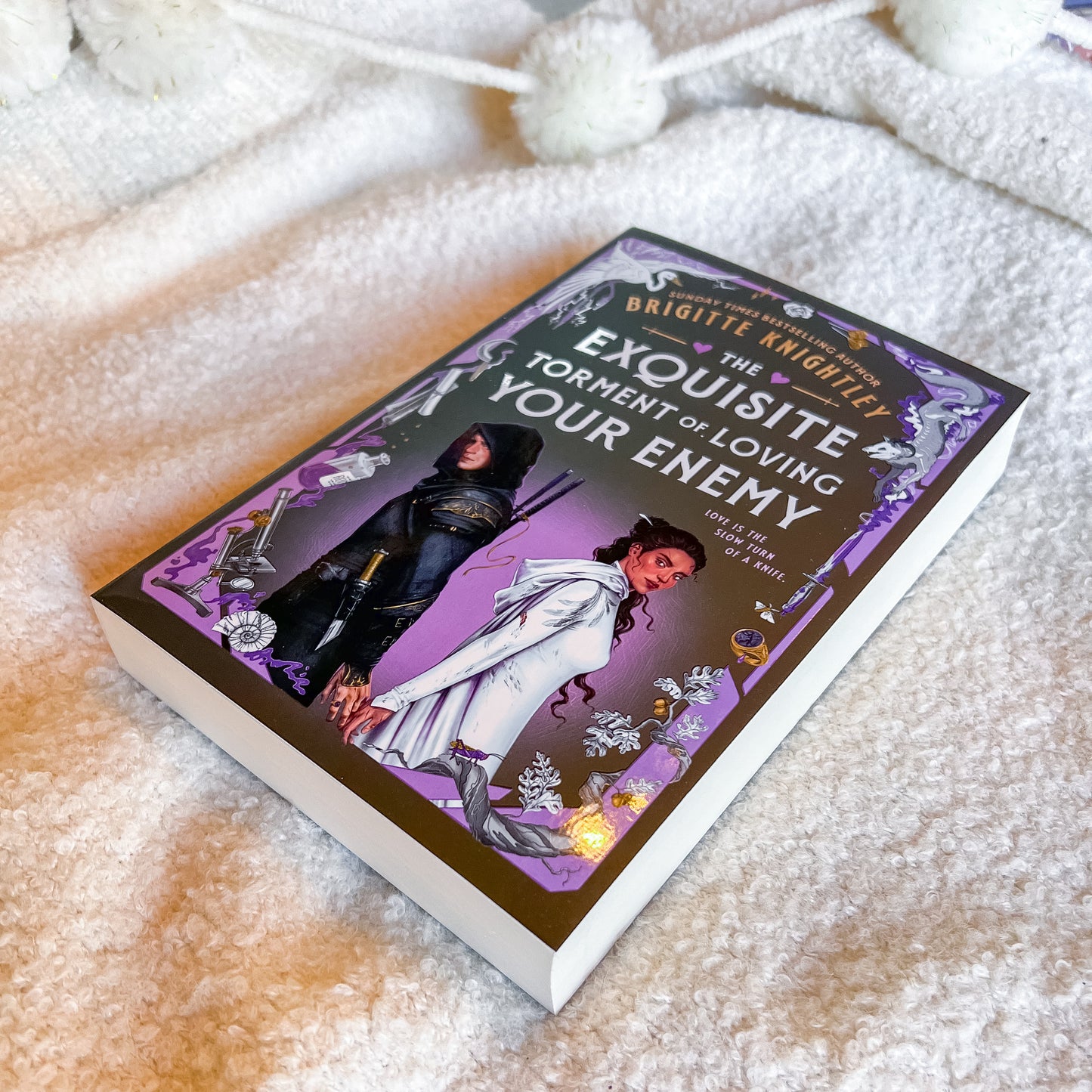 A hardcover book titled "The Exquisite Torment of Loving Your Enemy" by Brigittte Knobelitz lies on a soft, textured cream-colored blanket. The book cover features fantasy artwork with a purple background, a figure in white, and another in dark armor, with white pom-pom tassels visible at the top of the frame.