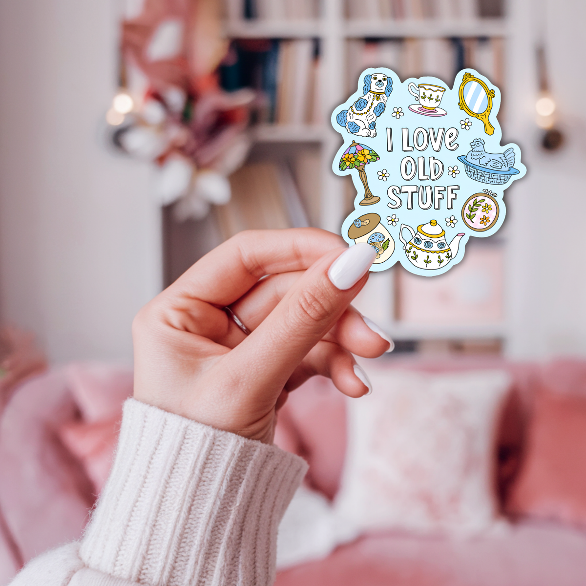 A hand with white manicured nails holds a die-cut sticker featuring retro tea-time illustrations and the text 'I LOVE OLD STUFF'. The background shows a cozy, softly blurred pink bedroom setting with a bed and shelves.