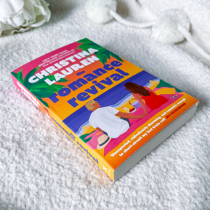 A paperback copy of the book 'The Romance Revival' by Christina Lauren lies on a textured white blanket, with white decorative flowers in the background.