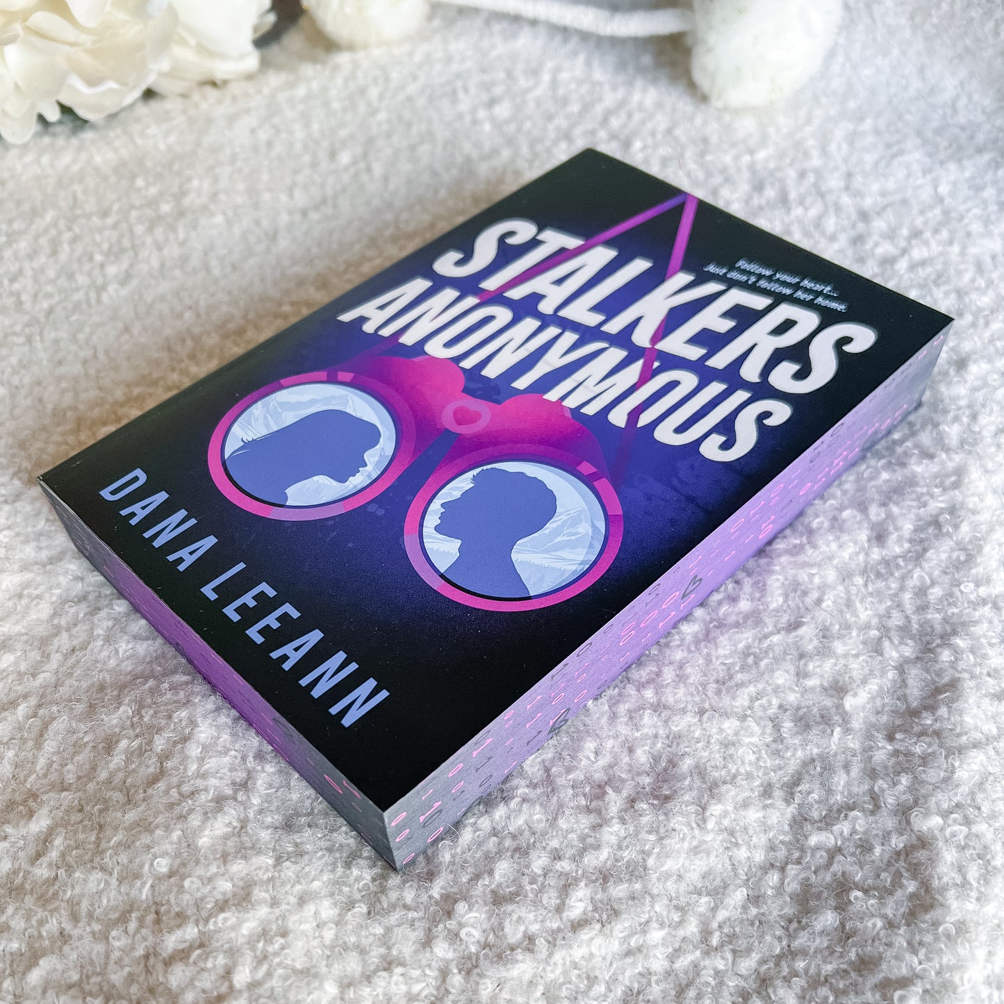 The book 'Stalkers Anonymous' by Dana Leeann, featuring a black cover with pink and purple sunglasses and white title text, resting on a textured white blanket.